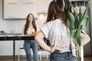 Mother sitting and daughter holding pink mother's day flowers and a hand-made card behind her back. The cover image for the Mother`s Day Collection.