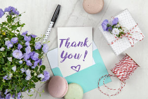 A table with craft supplies, a wrapped gift, a thank you card, loose macarons, and flowers. The cover image for the Thank You Collection.