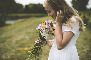 Woman dressed in white standing in a field holding and smelling a bouquet of freshly picked flowers. The cover image for the Get Well Collection.