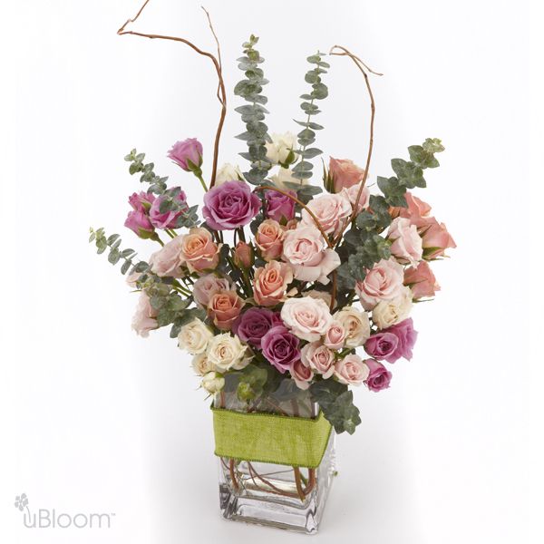 Floral arrangement with varying colors of spray rose, surrounded with eucalyptus and curly willow. The arrangement is placed in a tall cube vase, and a green grass ribbon