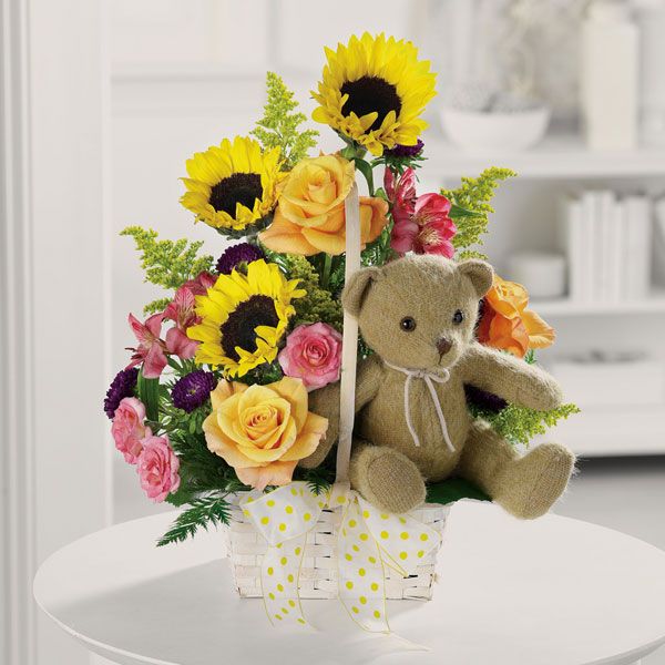 Floral arrangement on a weaved basket featuring roses, sunflowers and alstromeria, and finished with a nested teddy bear