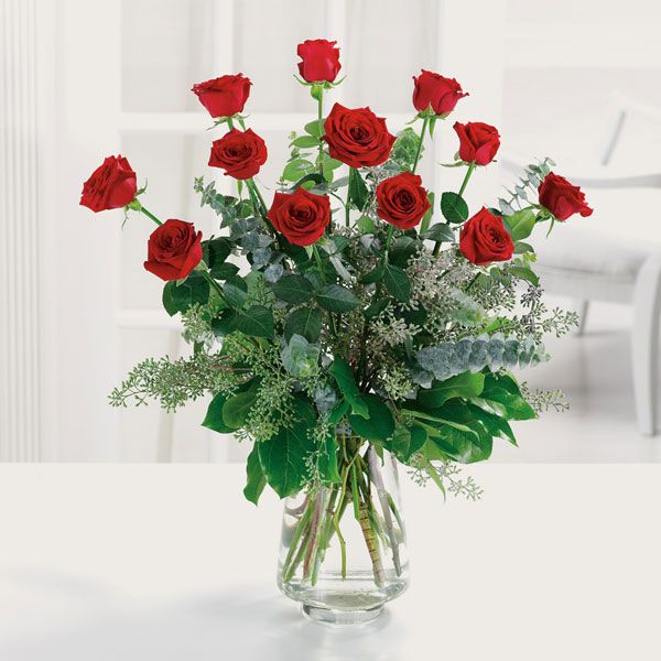 Clear glass vase with a dozen red roses and adorned by eucalyptus and other greenery.