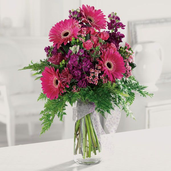 A pink and purple bouquet in a glass vase featuring several kinds of flowers, greenery and finished with an elegant ribbon