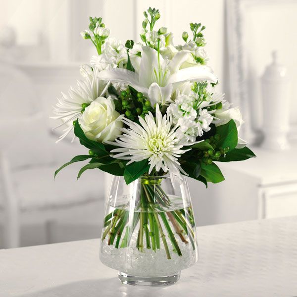 Floral arrangement in a clear glass vase featuring fragrant white Oriental lily surrounded by roses, Fuji mums, and white stock against a backdrop of lush greenery