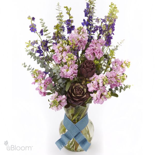 Ocean Side floral arrangement featuring larkspur, stock, artichoke, and eucalyptus in a clear glass vase with a blue ribbon.